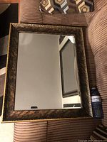 Full view of rectangular wall mirror with a broad, black and gold ornate frame resting on a textured tan fabric surface. Mirror reflects a partial wall and picture frame.