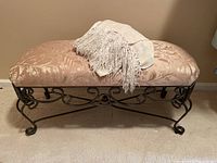 Front view of upholstery bench topped with folded off-white fringe throw blanket, showing pink damask fabric on seat and scrollwork metal base.