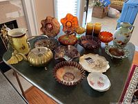 Full assortment of items on table including carnival glass, brass kettle, porcelain pitcher, plates, and candlesticks.