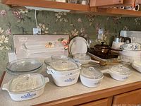 Photo showing assortment of CorningWare blue cornflower covered casseroles, clear glass bowls, and boxed Gallery Baker rectangular baker on a kitchen counter with floral wallpaper background.