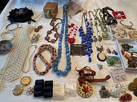Wide view of assortment of costume jewelry on white fabric background including multiple necklaces, a small jewelry box, bracelets, earrings, pins, and a black drawstring pouch