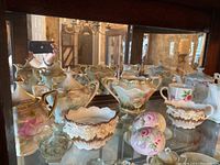 Porcelain sugar bowls, creamers and decorative eggs with handpainted floral designs and gold accents, displayed in a glass curio cabinet