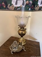 Full view of brass kerosene oil lamp and ornate silver-tone inkwell on wooden table, with wall tapestry in background.