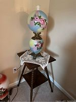 View of vintage painted glass globe lamp with roses on a wooden side table with crochet doily.
