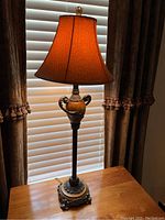 Single lamp on wooden surface next to window with blinds and dark curtains, lit amber shade showing warm lighting.