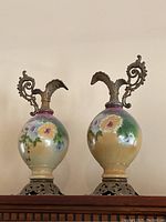 Front view of the pair of large round porcelain pitchers with detailed hand-painted floral designs and ornate metal spouts and scrollwork handles