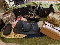 Wide shot showing assorted handbags and purses laid out on green bedspread, including a black leather handbag, a tan handbag, patterned purses, and a vintage box handbag with bird design.