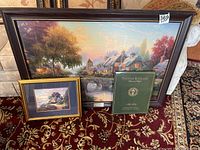 Three Thomas Kincaid prints on a carpeted floor, one large framed print of a village scene with a stone bridge, one smaller framed landscape print, and a green folder with another print.