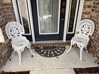 Two white metal bistro chairs placed outside on a concrete porch area against a brick wall and glass door.