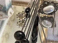 Close-up photos of assorted decorative hat pins with heads in black beads, floral patterns, and pearl-like designs roughly placed on a reflective surface.