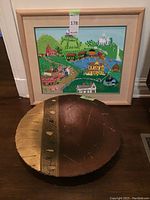 Framed colorful village painting by Marlene Zimmerman with a decorative plate featuring brown and gold geometric textures in front.