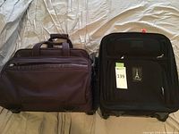 Two carryon suitcases side by side, one black with multiple front pockets, one dark purple soft-sided suitcase.
