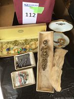 All jewelry pieces and porcelain box in view: gold-tone set in yellow box, rhinestone earrings card, beaded bracelets bag, filigree brooch, porcelain box