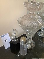 Photo showing large crystal decanter with stopper, salt shaker with silver top, and crystal mushroom figurines on black pedestal base.