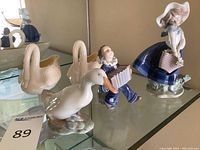 Photo of Royal Copenhagen swan pitcher, Lladro boy figurine, and Belleek girl figurine on glass shelf