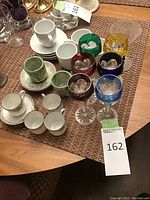 Top view of the set showing multiple ceramic tea cups, saucers, coffee mugs, and colored stemware goblets in various colors arranged on a woven placemat.