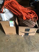 Two cardboard boxes on ground, one containing orange heavy-duty extension cords and the other containing paper bags and miscellaneous cables.