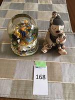 Overall shot showing both the Hanukkah-themed snow globe with three children figurines inside and Native American storyteller figurine by Anita F. Toya on a tablecloth.