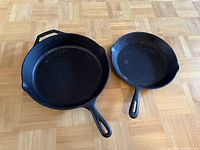 Two cast iron skillets side by side on wooden floor showing overall condition and size difference.