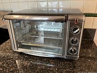 Front angle view of the Black+Decker toaster oven showing stainless steel exterior, glass door, metal handle, and interior wire rack and baking tray.
