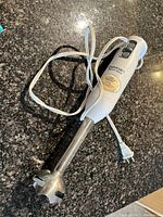 Full view of the hand blender showing white plastic body with attached power cord and stainless steel blending shaft.