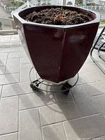 Front view of the glazed planter urn showing the octagonal shape, deep burgundy glaze, soil inside, and wheeled metal base.