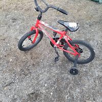 Red Huffy Rock It boys' bicycle with training wheels on gravel surface.