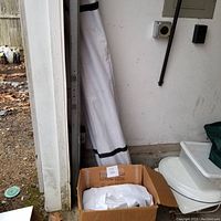 White rolled canopy tent frame leaning against wall behind tent fabric in a cardboard box.