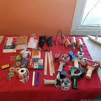 Full overview of office supply items laid on red cloth, including tape dispensers, rulers, lint rollers, receipt pads, lighters, label rolls, and other accessories