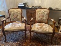 Front view of two matching carved wood armchairs with floral tapestry upholstery