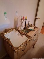 Front and top view of wood dresser showing three drawers with floral details and large ornate gold-framed mirror on top along with other decorative items.