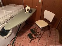 Wide shot of GE iron on ironing board next to retro chair and step stool in basement room with pink tile floor and wood panel walls