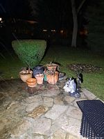 Garden patio scene at night. Shows collection of orange and blue garden planters with dried plants. Figurines of a white rabbit, blue owl with large eyes and two tall dark birds are displayed on stone patio.