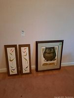 Three framed pieces leaning against a wall: two narrow bird prints in wood frames and one larger owl print in a dark frame