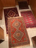 Four small rugs laid out on tile floor showing orange medallion rug at front, geometric runner at back left, red boteh rug at back right and dark floral rug at top right.