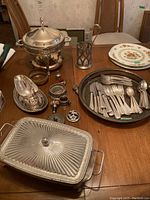 Silver plated cutlery spread on a table with serving dishes, bowls, gravy boat, silver trays, and plates in background