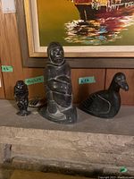 Three soapstone carvings displayed together including small signed piece on left, tall Inuit man in center, goose figurine on right