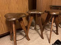 Three matching bar stools with triangular brown leather seats and wooden legs, showing nailhead trim and metal footrest rings, one seat with wear.