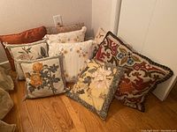 Lot of 9 decorative pillows on wooden floor showing variety of patterns and textures.