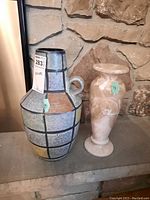Front view of both vases placed side by side on a stone surface in front of a stone wall showing relative size and colors