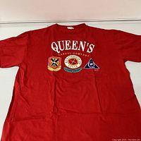 Full front view of red Queens University shirt with printed text and patches showing three university-related logos.