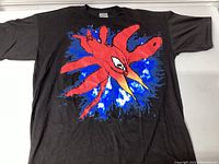 Front view of black T-shirt with large colorful abstract graphic design of a red bird-like figure on blue splash.