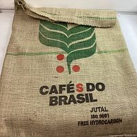 Front side of burlap coffee bean bag showing green coffee leaves graphic, red coffee cherries, and black text 'CAFÉS DO BRASIL' with additional text 'JUTAL ISO 9001 FREE HYDROCARBON'. Material is coarse woven jute with frayed top opening.