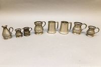 Full view of all 8 pewter mugs and pitcher lined up on plain background to show variety and approximate sizes