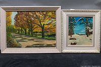Two framed oil on board paintings by Winston Thompson displayed side by side. Left painting shows an autumn trail among trees. Right painting shows a snow-covered forest scene.