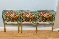Three vintage metal TV trays leaning against wall, showing floral and fruit still life designs on tray tops and foldable gold-toned metal legs.