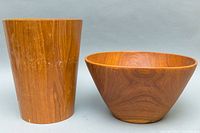 Photo showing two wooden items, a large bowl and a tall cylindrical basket, both made of teak wood and placed side by side on a gray background.