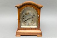 Front view of wooden Junghans mantle clock with curved top and glass door closed showing aged metal dial with Arabic numbers.