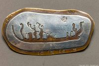 Front view of brooch showing engraved Native American style humanoid figures and sun design on an irregular oval bronze base with a silver-tone metal plaque.