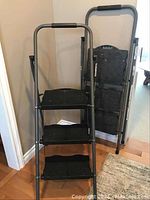 Two Cosco 3-step ladders, one opened showing steps and hand bar, the other folded against the wall.